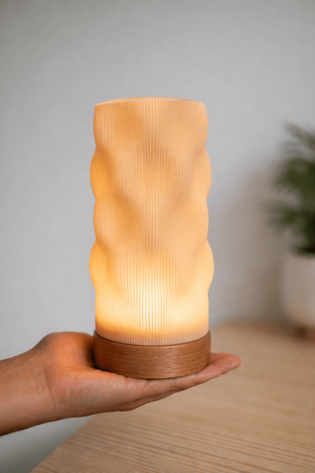 Wavia bordlampe tændt med varmt gult lys, holdt i hånd – organisk bølgeform i beige PLA